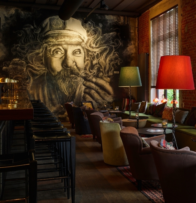 A cozy bar lounge with patterned cushions, round tables, colorful floor lamps, and large mural art of bearded face on the wall.