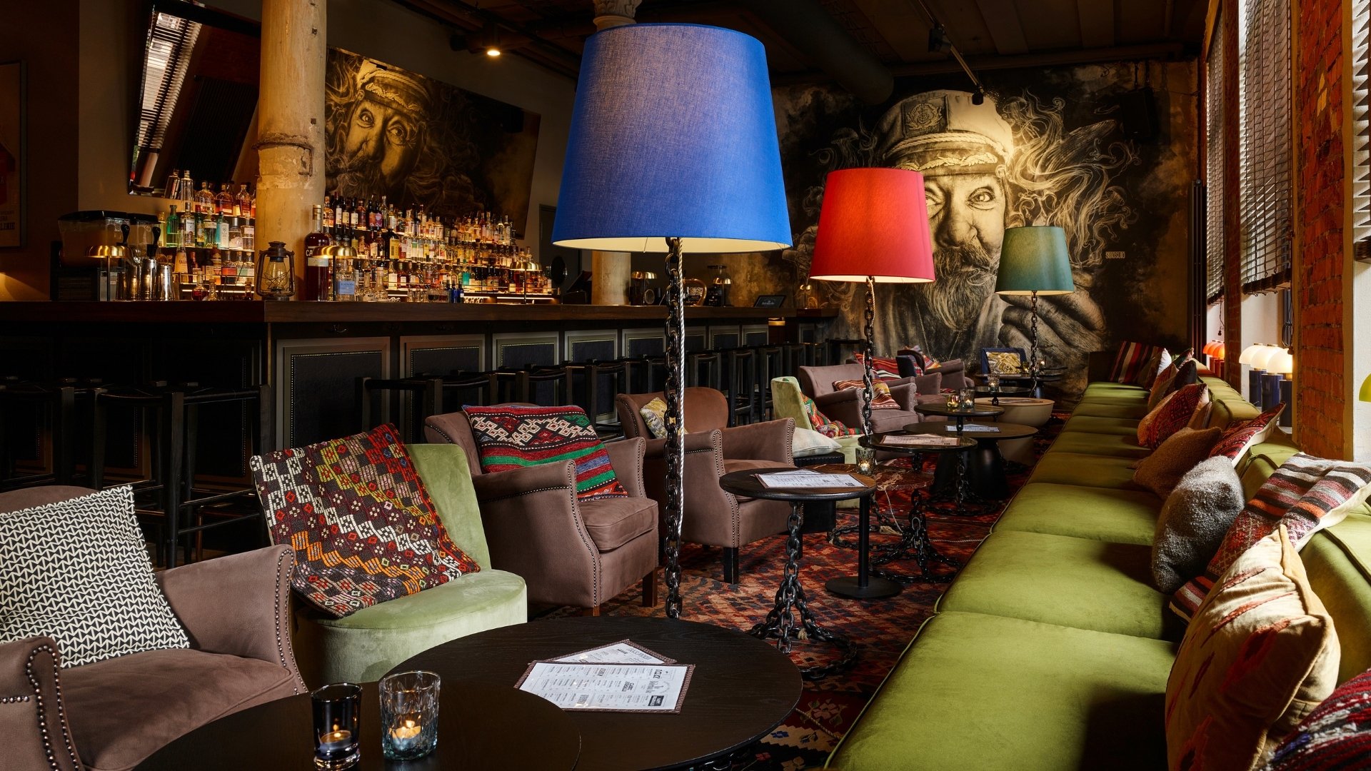 A cozy bar lounge with patterned cushions, round tables, colorful floor lamps, and large mural art of bearded face on the wall. Bottles line the shelves behind the bar counter.