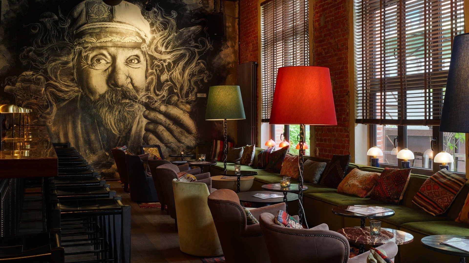 A cozy bar lounge with patterned cushions, round tables, colorful floor lamps, and large mural art of bearded face on the wall. Bottles line the shelves behind the bar counter.