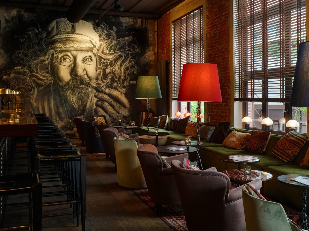 A cozy bar lounge with patterned cushions, round tables, colorful floor lamps, and large mural art of bearded face on the wall.