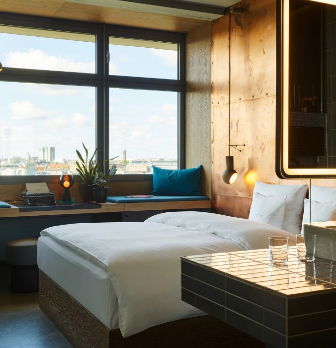 Modern hotel room with a cosy bed and wooden details. A large window offers a city view, with a plant and blue cushions on the windowsill, creating a serene atmosphere.