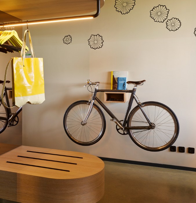 Hotel room featuring a wall-mounted bicycle, a yellow tote bag, and books on a shelf, with flower decals on the wall.
