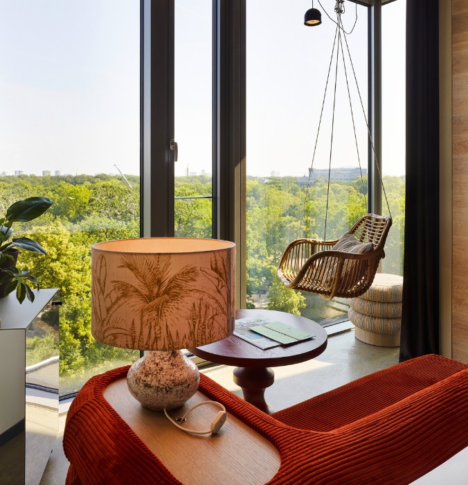 Hotel room featuring a woven swing chair by large windows, a palm-patterned lamp, and lush greenery outside.