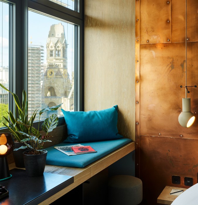 Window seat with a teal cushion and plant, overlooking a cityscape featuring a historic tower through large windows. Warm, modern decor.