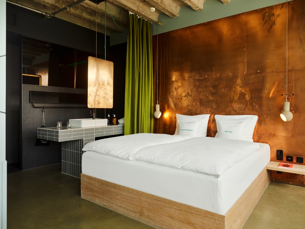 Modern hotel room featuring a double bed, copper wall accents, green curtains, and a bathroom area.