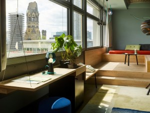 Hotel room with large windows showing a cityscape, including an old church. The space features a desk with plants and a lamp, and a red couch on wooden steps.