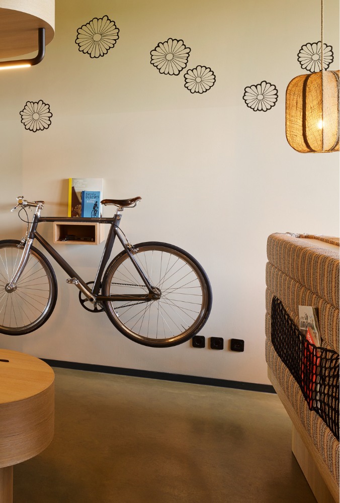 A minimalist room with a bike mounted on a white wall, featuring black floral decals. A pendant light hangs beside a wicker-patterned chair, creating a cosy vibe.