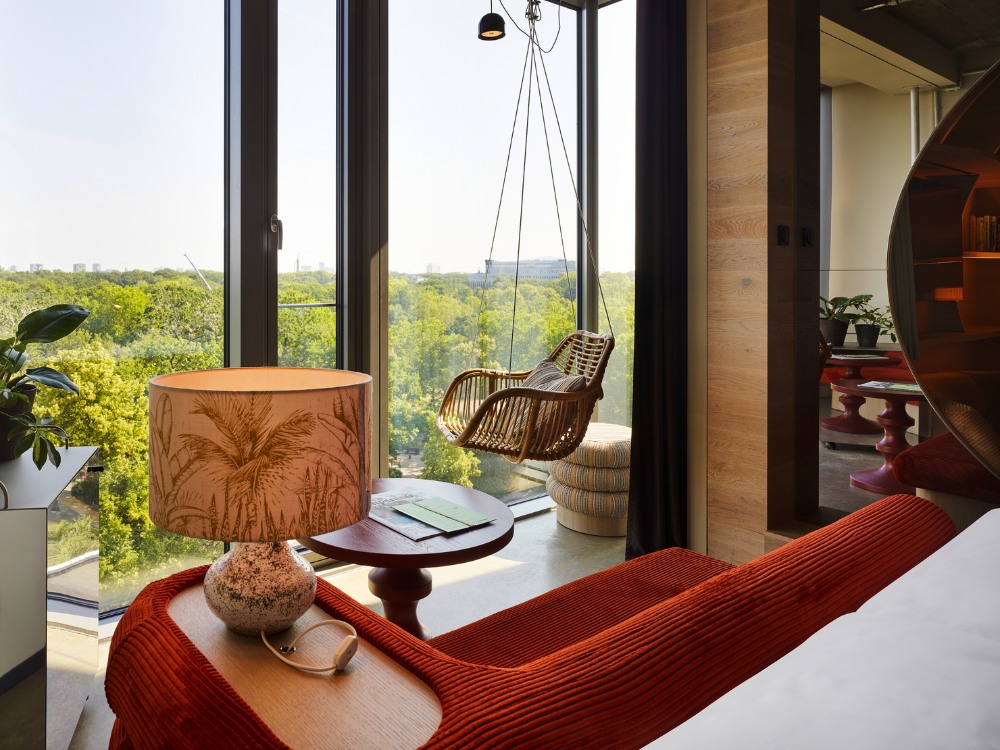 Hotel room with large windows showing a lush green view. Features a rattan swing chair, red sofa, round side table, and decorative lamp. Relaxed ambiance.
