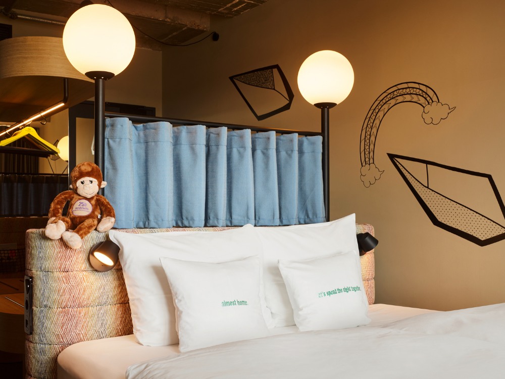 Bed with white pillows, one reading "almost home," and a plush monkey sits on a checkered headboard. Warm lights illuminate a whimsical mural.