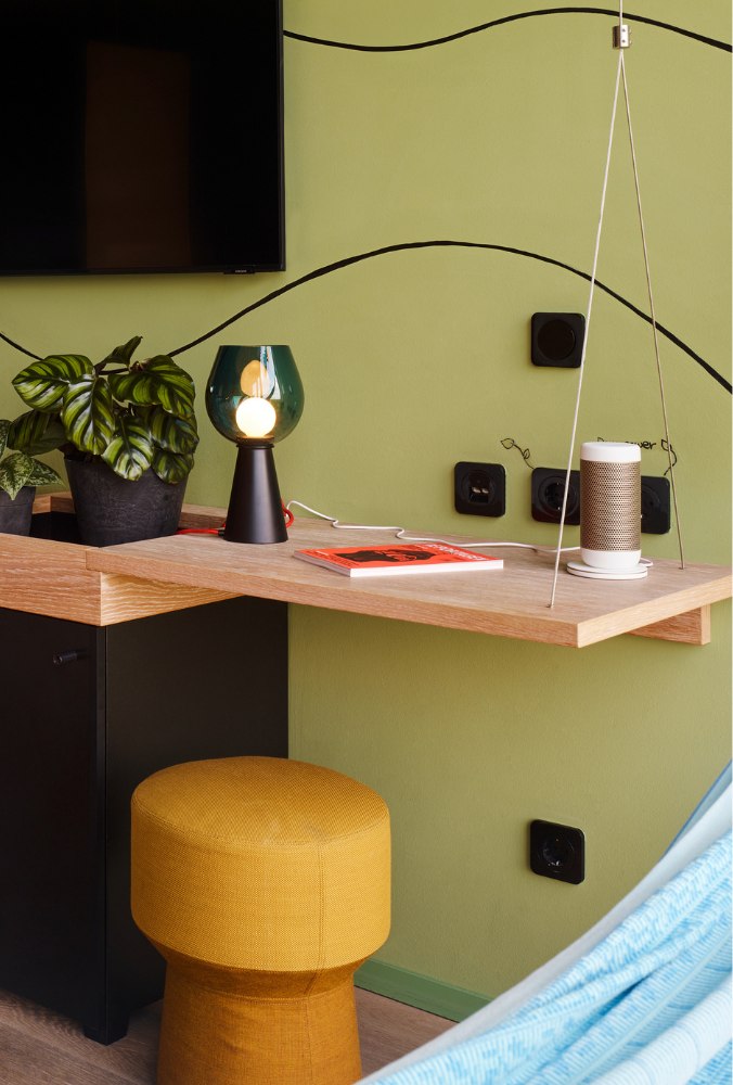 A modern interior scene with a wooden desk against a light green wall. It features a potted plant, lamp, book, and speaker. Yellow stool adds a pop of color.