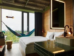 Hotel room with a large window showcasing a hammock and a view of green trees. Minimalistic decor includes a bed, illuminated mirror, and stylish lighting.