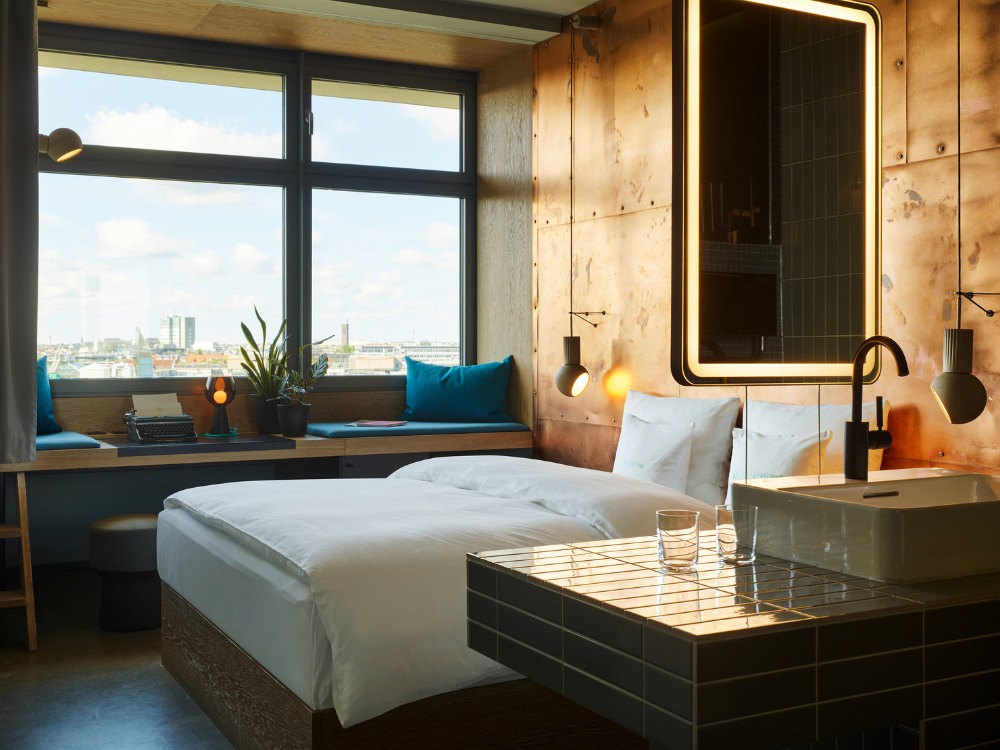 Hotel room with a double bed, large windows, a workspace, and stylish decor featuring a metallic accent wall and sleek sink.
