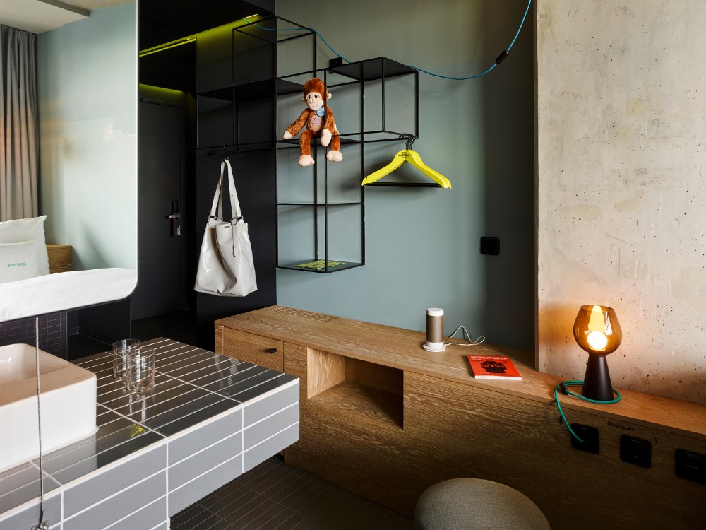 Hotel room featuring a monkey cuddly toy, green accents, modern furniture, and a warm lamp on a wooden shelf.