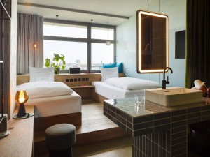 Hotel room featuring two beds, a modern sink, and large windows with city views, accented by soft lighting and greenery.