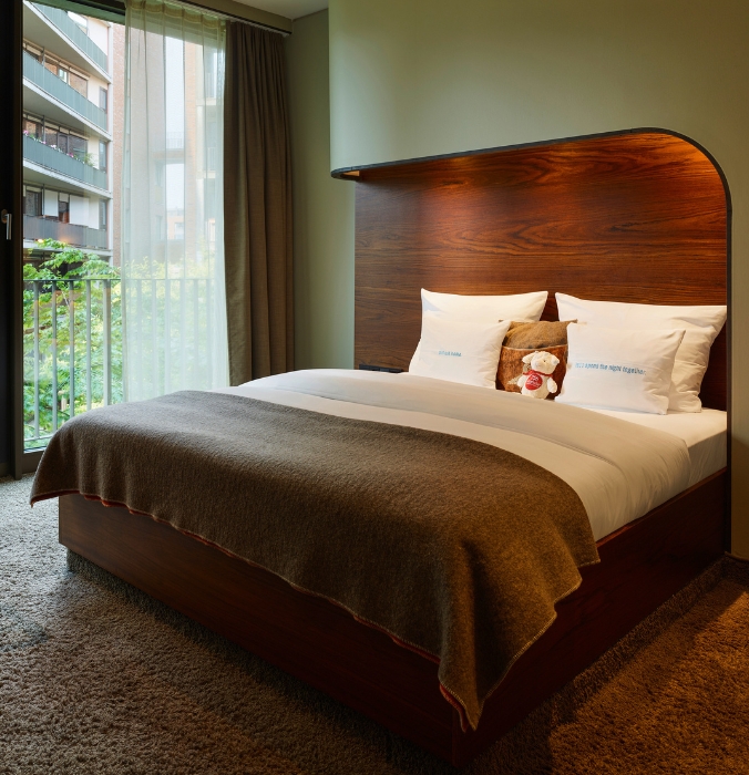 Hotel room with a wooden bed, cosy blankets, plush carpet, and large curtains revealing a view outside.