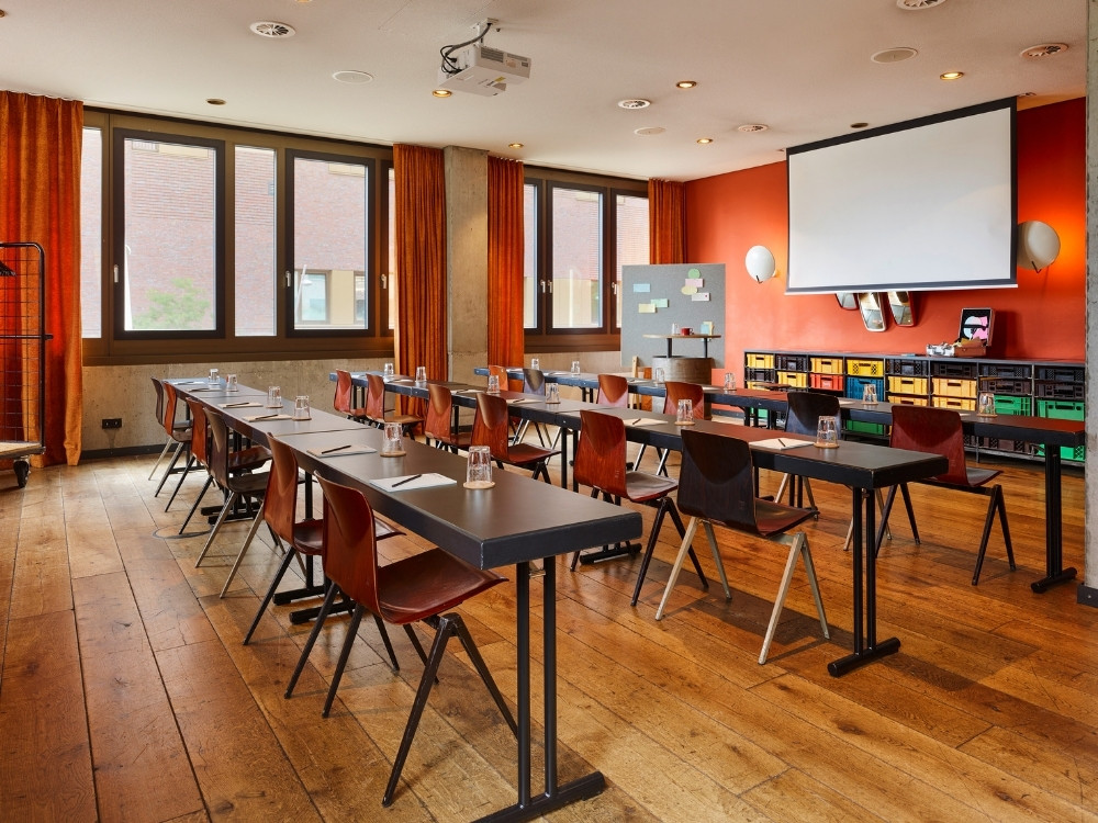 Modern meeting room with wooden floors, black tables, chairs, projector screen, and large windows, set up for a presentation.