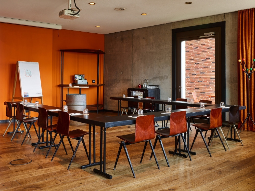 Meeting room with tables and chairs arranged for a presentation, featuring an orange wall, projector, and a door to an outdoor space.