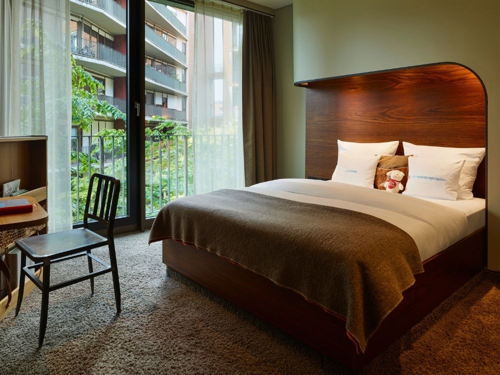 Hotel room with a wooden bed, cosy blankets, plush carpet, and large curtains revealing a city view outside.