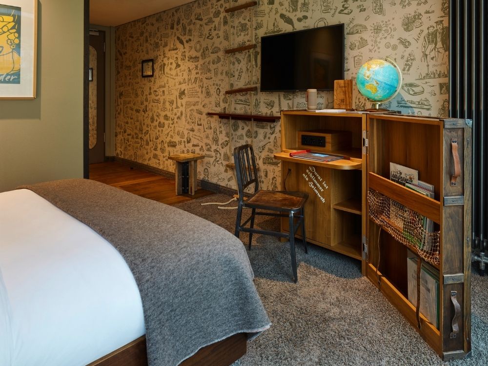 Hotel room with a bed, gray blanket, wooden desk and chair, open cabinet with a globe, wall-mounted TV, and illustrated wallpaper.
