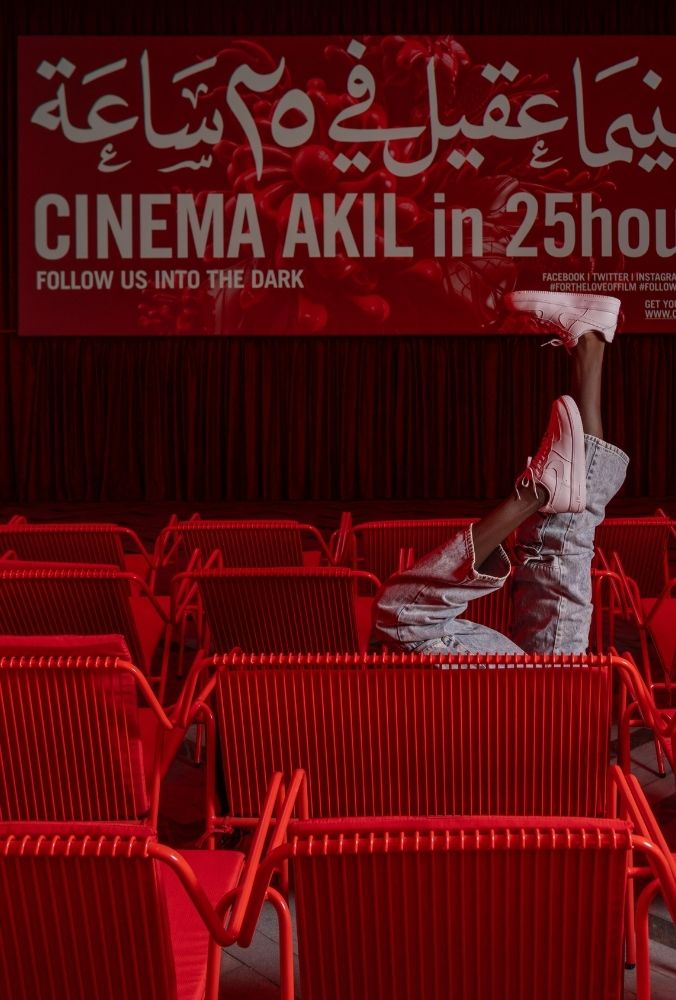 A person kicks their feet playfully wearing pink sneakers and jeans among rows of empty red chairs. A bold banner reads "CINEMA AKIL in 25hours."