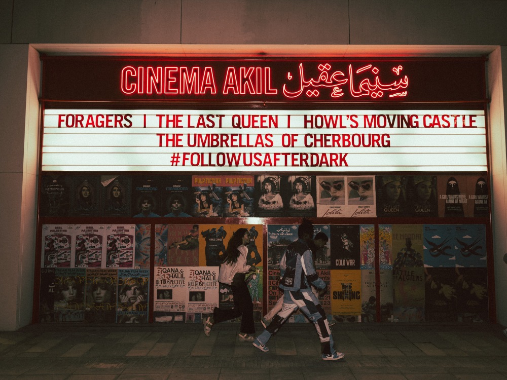 Marquee of "Cinema Akil" glowing in red at night, showcasing films like "Howl's Moving Castle." People walk past, creating a lively, urban atmosphere.