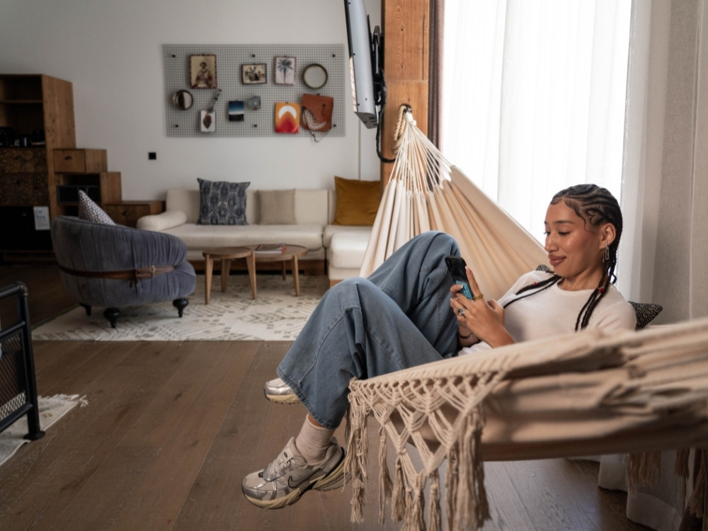 A person relaxes in a hammock, using a phone, surrounded by a stylish, modern living room with a cosy couch and decorative wall art.