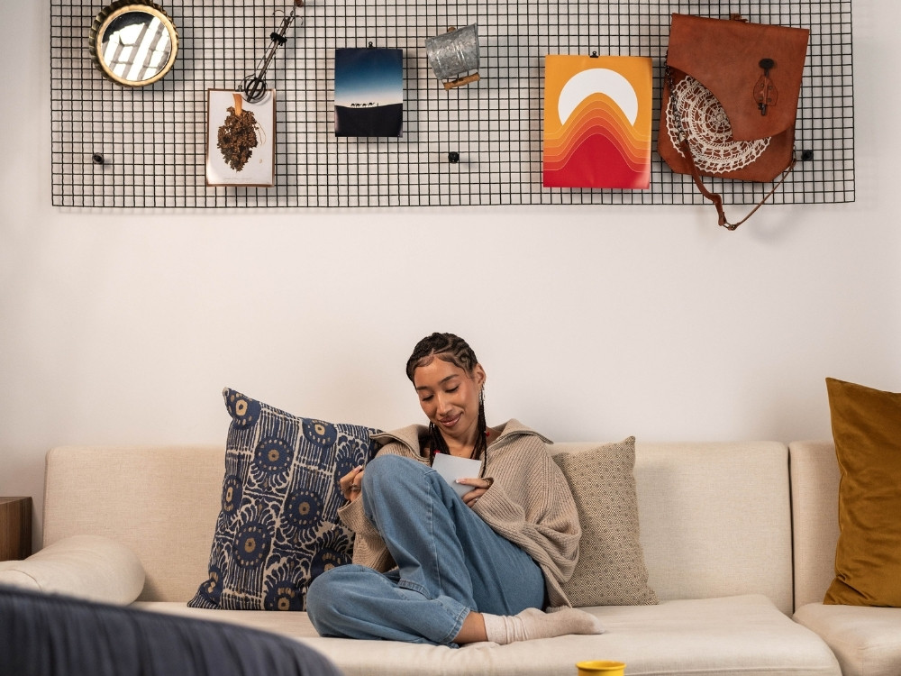 A person sits comfortably on a beige couch, casually dressed, with decorative wall hangings and pillows accentuating the cosy living space.