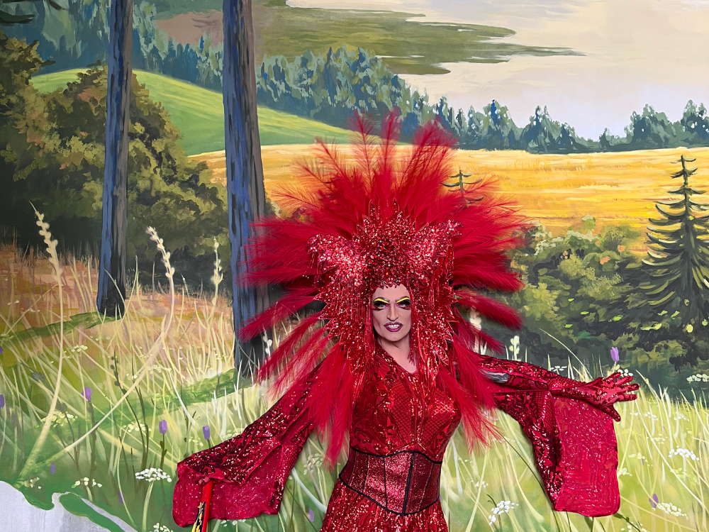 Drag queen in a sparkling red outfit with dramatic feathered headpiece, standing in front of a vibrant forest and meadow mural, smiling with arms open wide.