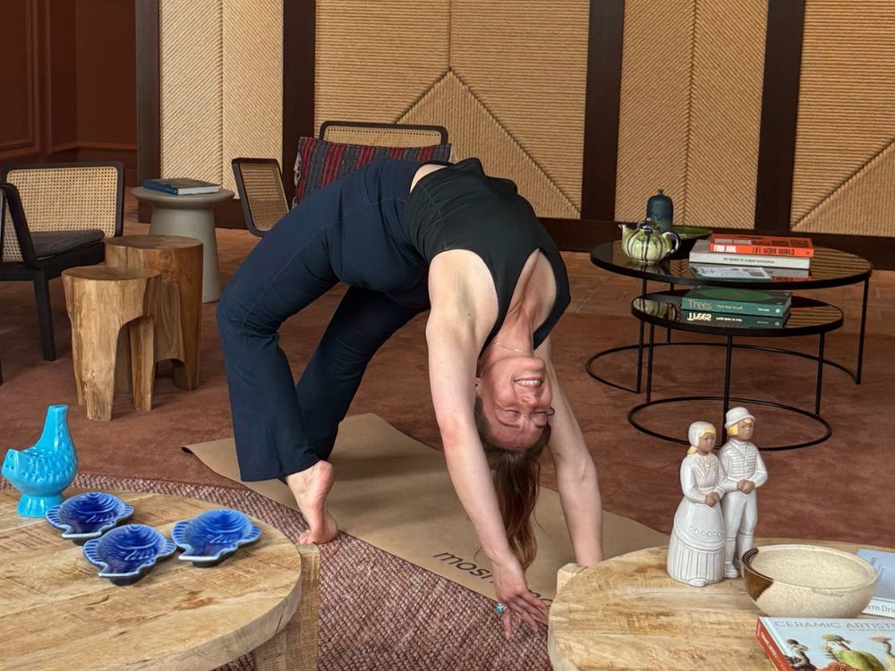ChatGPT said:A woman practices yoga indoors, performing a backbend on a mat with a joyful expression. She is surrounded by cozy, modern furniture, wooden tables, ceramics, and books, creating a warm and inviting atmosphere.