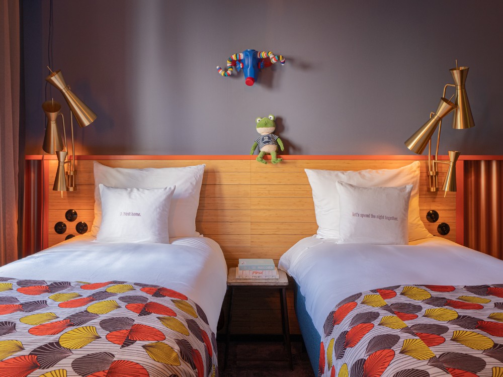 A cozy hotel room with two beds, colorful patterned comforters, stylish lamps, and a green stuffed frog on the headboard.