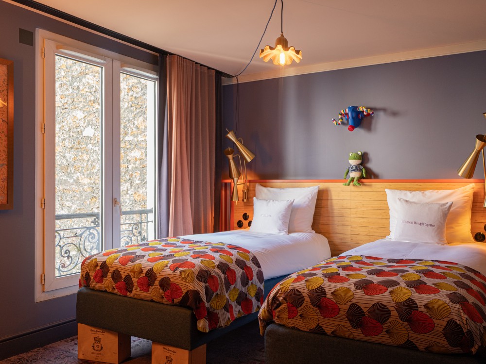 Brightly colored twin beds in a cozy hotel room with whimsical decor, featuring a playful elephant and frog plush toys. Natural light floods in.