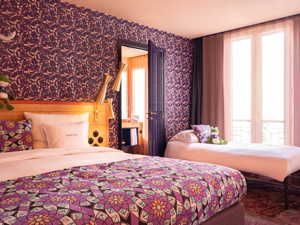 Stylish hotel room with colorful patterned bedding, a plush toy frog, and large windows offering natural light.