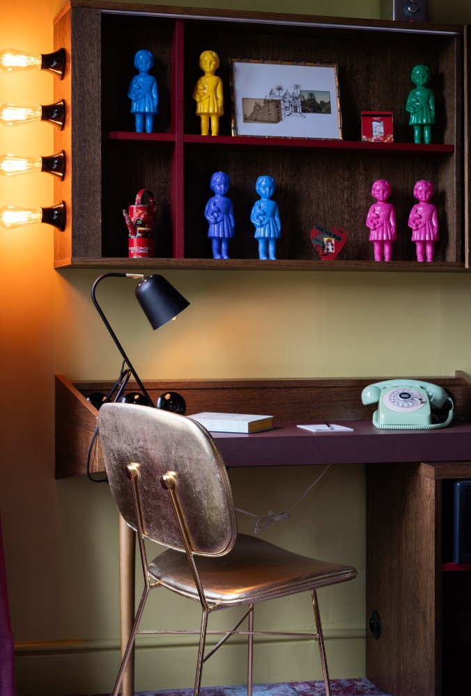 A colorful home office corner featuring a gold chair, vintage rotary phone, and a shelf displaying vibrant plastic dolls and an old photo.