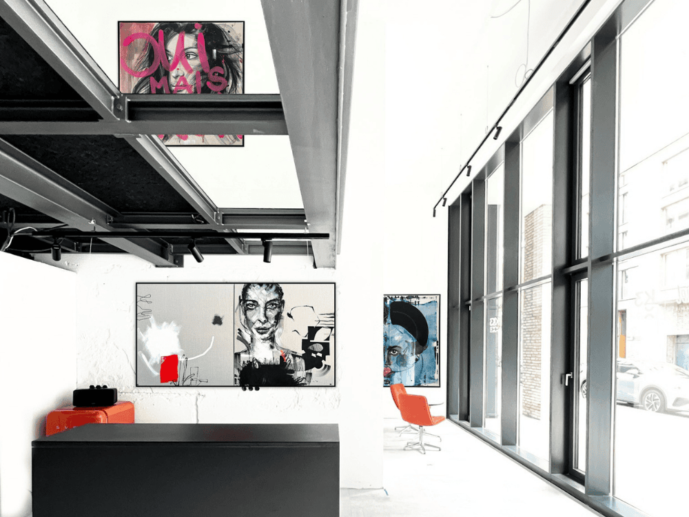 A modern art gallery featuring vibrant paintings, large windows, and contemporary furniture, with a striking orange chair in the corner.