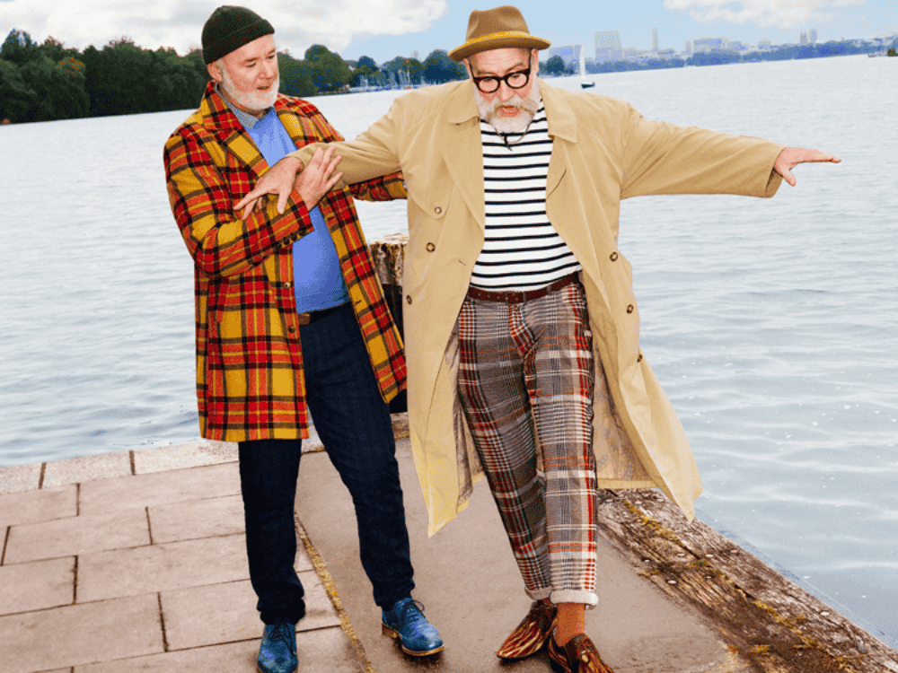 Two people stand near a calm lake, one playfully balancing on a wooden edge, dressed in colorful outfits and coats, surrounded by nature.