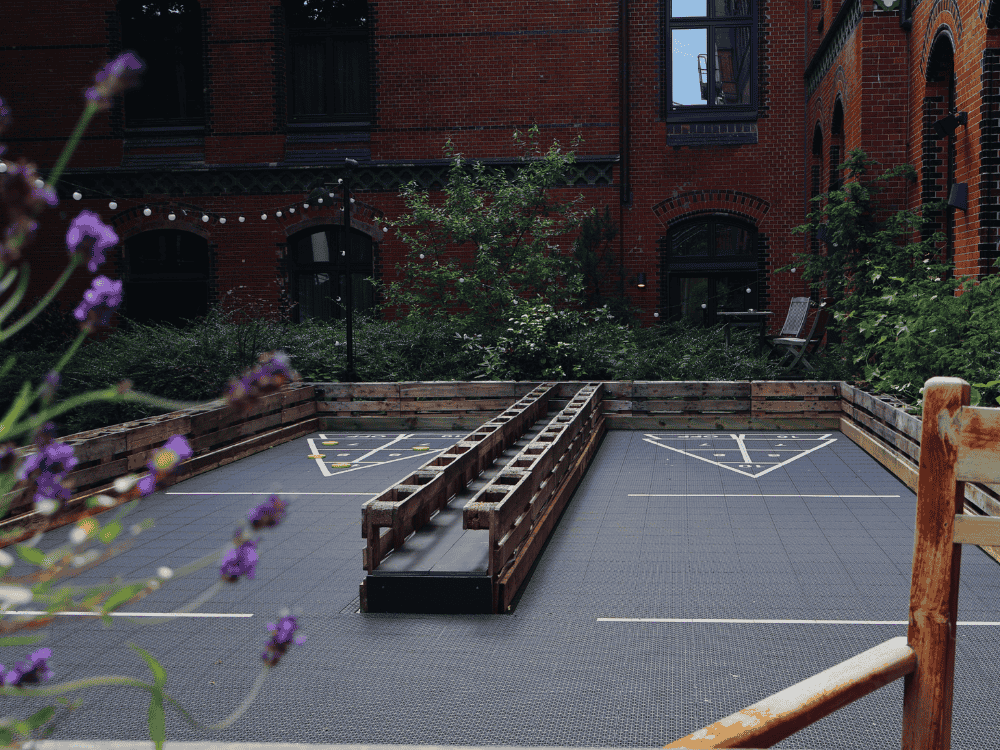 A wooden-bordered outdoor gaming area featuring a checkered floor, with a sloped lane for tossing objects, surrounded by greenery and brick walls.
