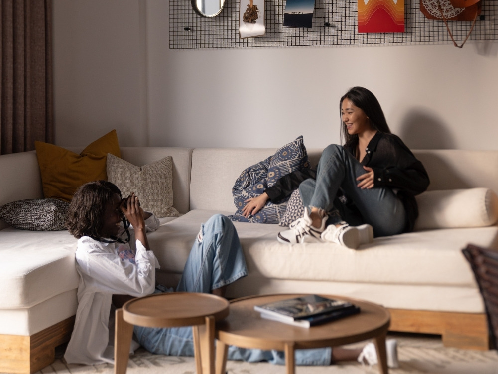 Two friends relax in a cozy living room. One sits on the sofa, smiling, while the other lounges on the floor, taking photos. Warm, inviting atmosphere.