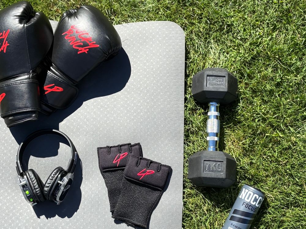 Outdoor workout setup on the grass with black Lucky Punch boxing gloves, wrist wraps, wireless headphones, a 7kg dumbbell, and a NOCCO energy drink placed on a grey exercise mat.