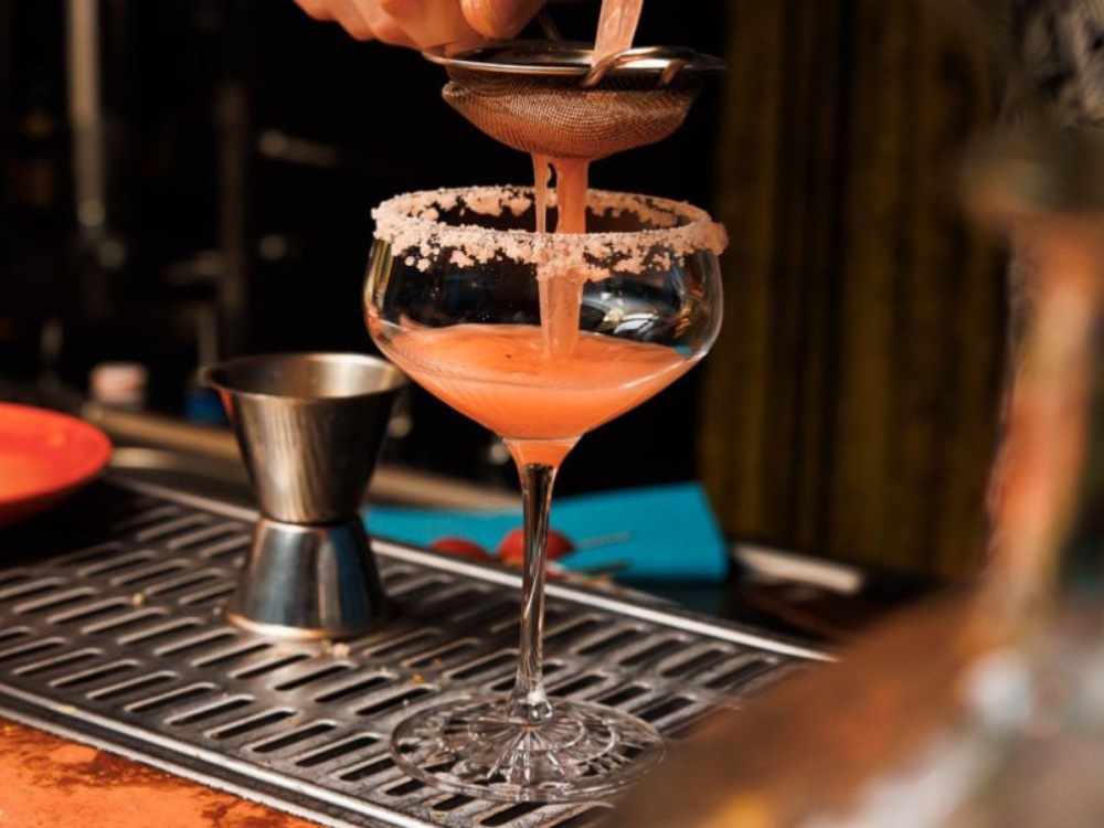 A bartender pours a pink cocktail through a strainer into a salt-rimmed glass on a bar counter, with a jigger in the background.