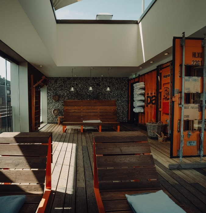 Modern sauna recreaation area with wooden deck chairs, bright windows, and an industrial-style wall featuring colorful shipping containers.