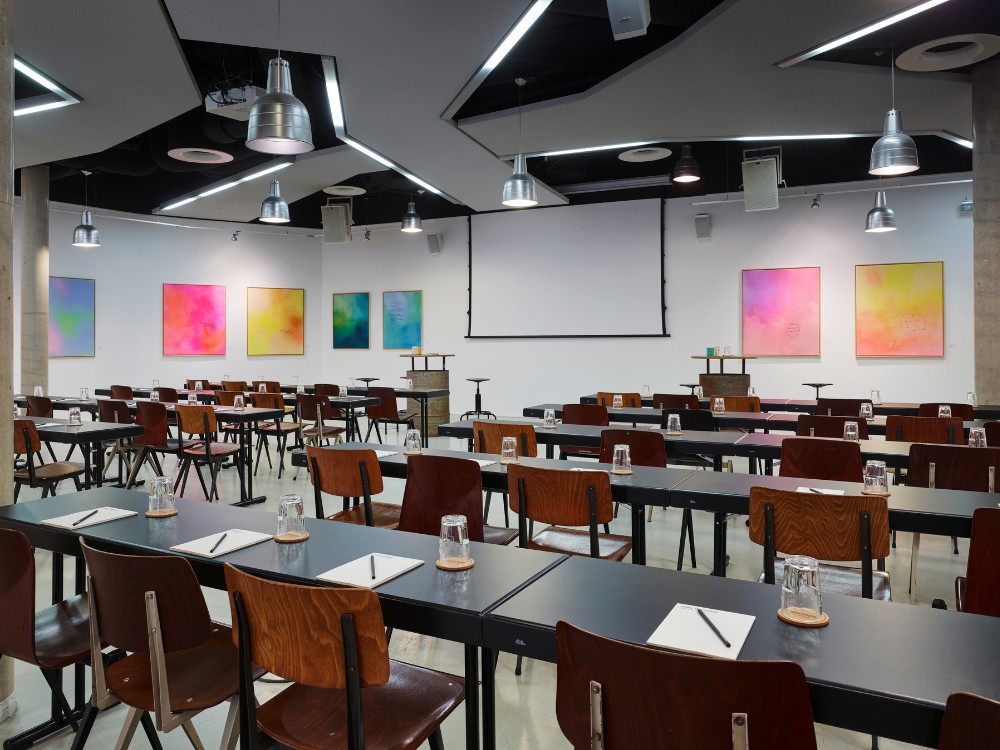 Modern conference room with rows of tables and chairs, colorful artwork on the walls, and a large screen at the front.