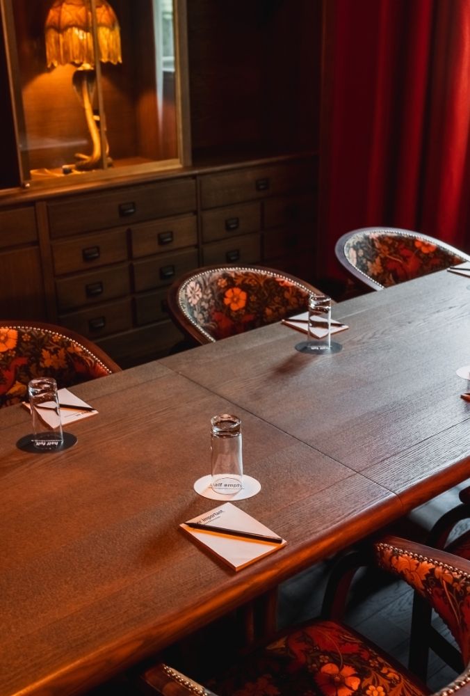 A wooden conference table with four floral upholstered chairs, each place set with a glass, a coaster, and a folded napkin. A lamp and drawers are in the background.