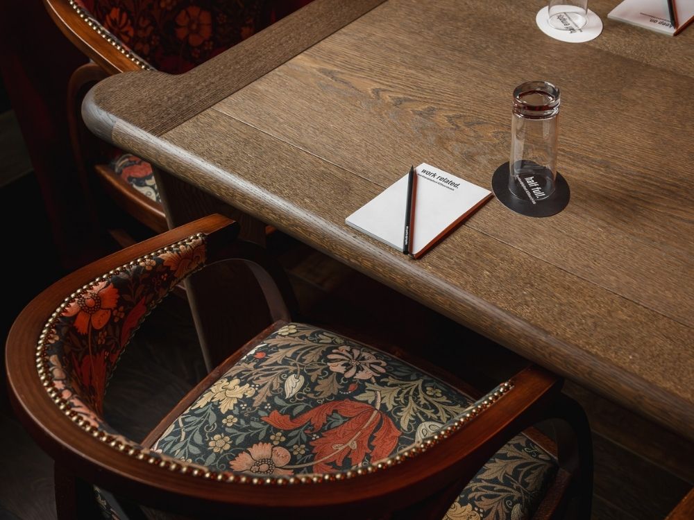 A wooden table with a notepad, pen, glass, and coaster next to a cushioned chair with a colorful floral pattern.