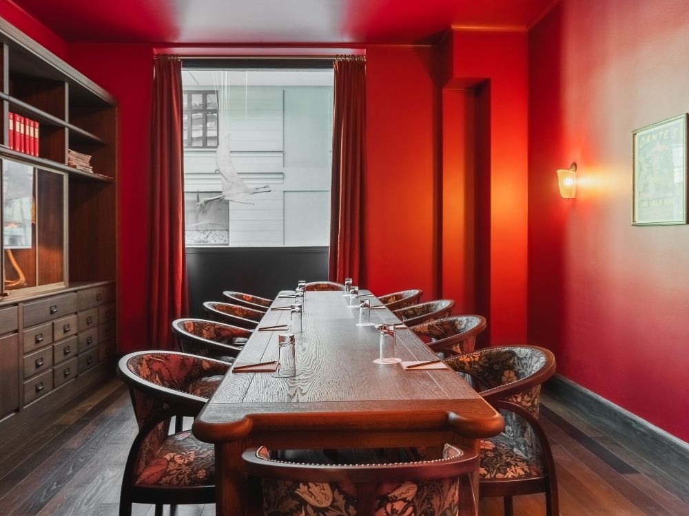 Elegant meeting room with deep red walls and curtains, a long wooden table set for ten, cushioned chairs, warm lighting, and a vintage ambiance.