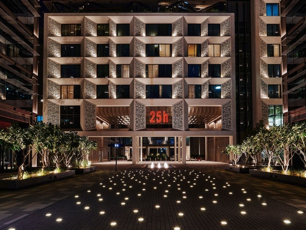 Modern hotel facade at night with geometric windows, warm lighting, and a glowing red "25h" sign. Foreground features lit pathway with trees.