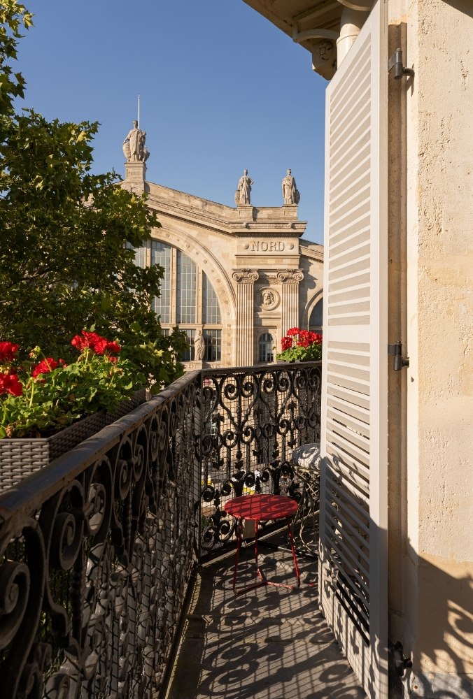 A sunlit balcony with red flowers and a small table overlooks the ornate architecture of the Gare du Nord train station.