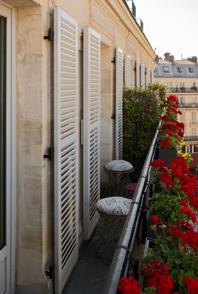 A cosy balcony with white shuttered windows, stylish stools, and vibrant red geraniums in decorative pots lining the railing.