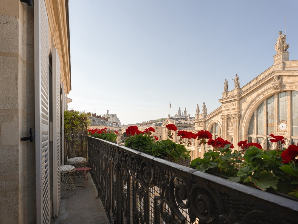 A sunlit balcony with red flowers and a small table overlooks the ornate architecture of the Gare du Nord train station.