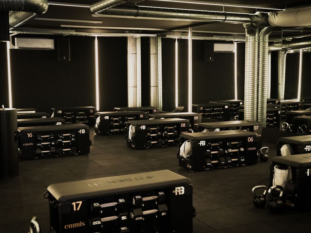 Dimly lit gym with black exercise stations, each numbered and equipped with weights and kettlebells. A sleek, modern atmosphere is emphasized by metallic ducts.