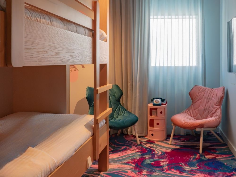 Cosy room with wooden bunk beds, two colorful cushioned chairs, a round side table with a rotary phone, and a vibrant patterned carpet.